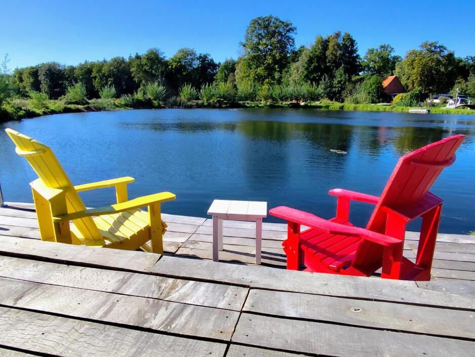 Bunte Adirondack-Stühle auf einem Holzsteg direkt am Mühlenteich der Wassermühle Bruchmühlen – ein idyllischer Ort zum Entspannen und Genießen der Natur.
