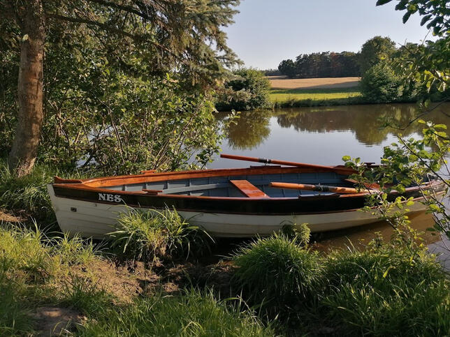 Ein traditionelles Ruderboot liegt am Ufer eines ruhigen Sees, umgeben von Wiesen, Bäumen und einer idyllischen Naturlandschaft.