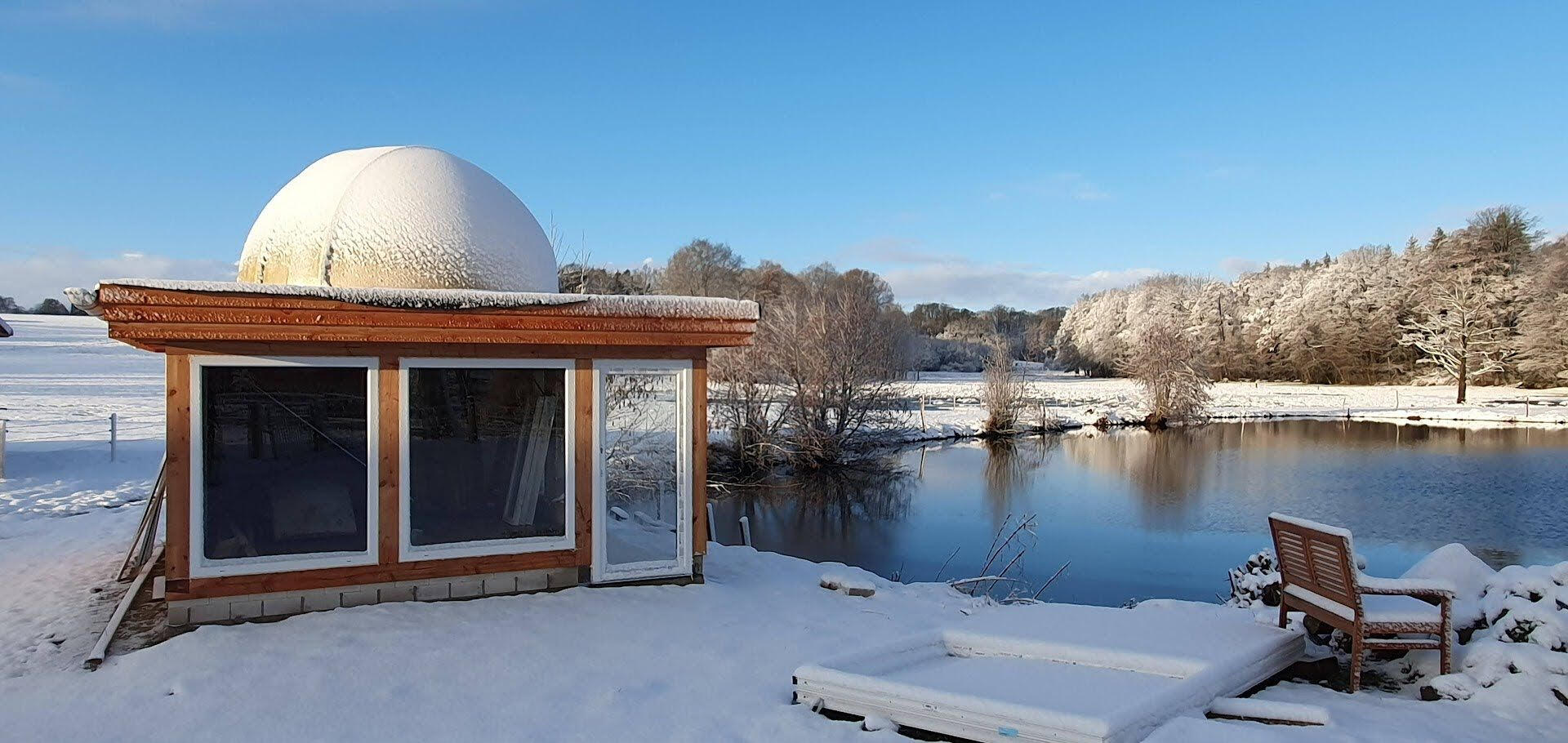Verschneite Sternwarte mit Kuppel direkt am See an einem sonnigen Wintertag – ein idyllischer Ort für Astronomie und Naturerlebnisse.